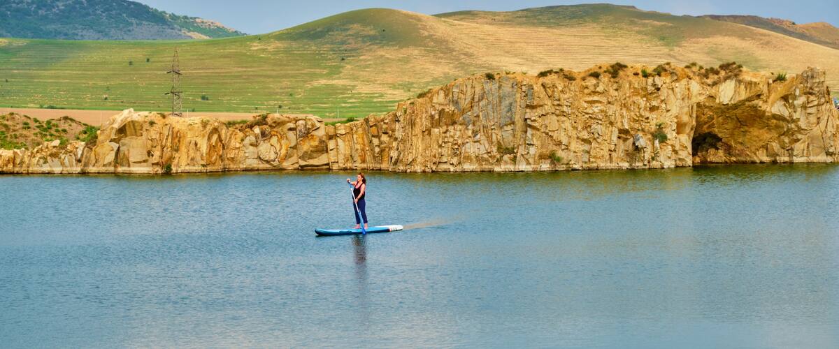 Young female tourist paddle-boarding on an isolated lake, with space for text at the bottom. Iacobdeal lake in Turcoaia, Tulcea, Romania at sunset, with warm light.