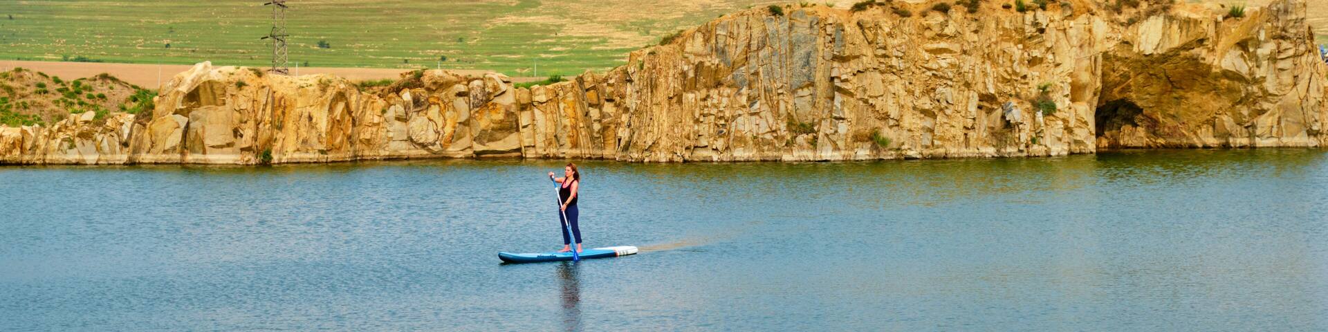 A person paddleboarding on a calm lake with a rocky cliff and hills in the background.