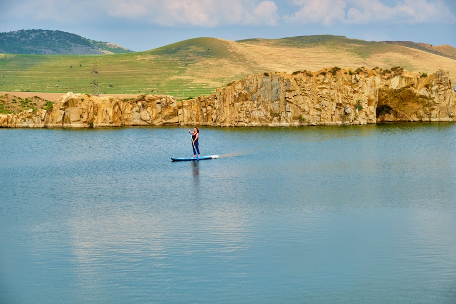 Uma pessoa praticando paddleboard num lago calmo, com um penhasco rochoso e colinas ao fundo.