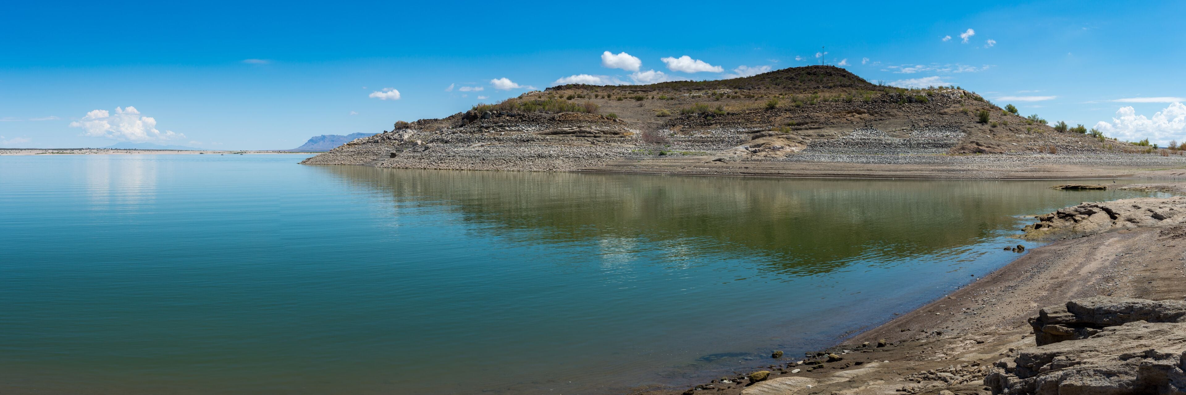 A calm lake with a rocky shoreline and a hill in the background.