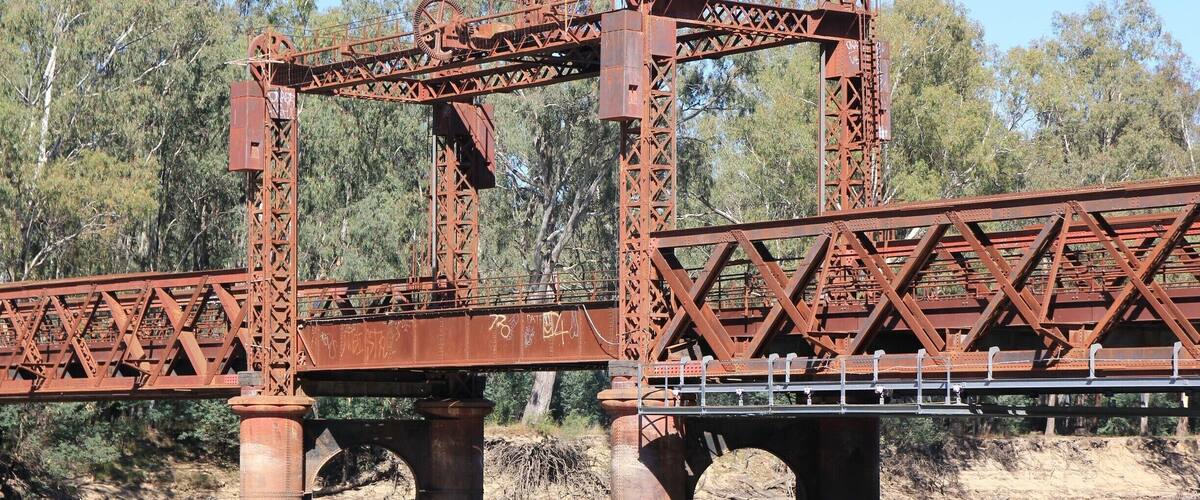 A rusted metal bridge over a river with trees in the background.