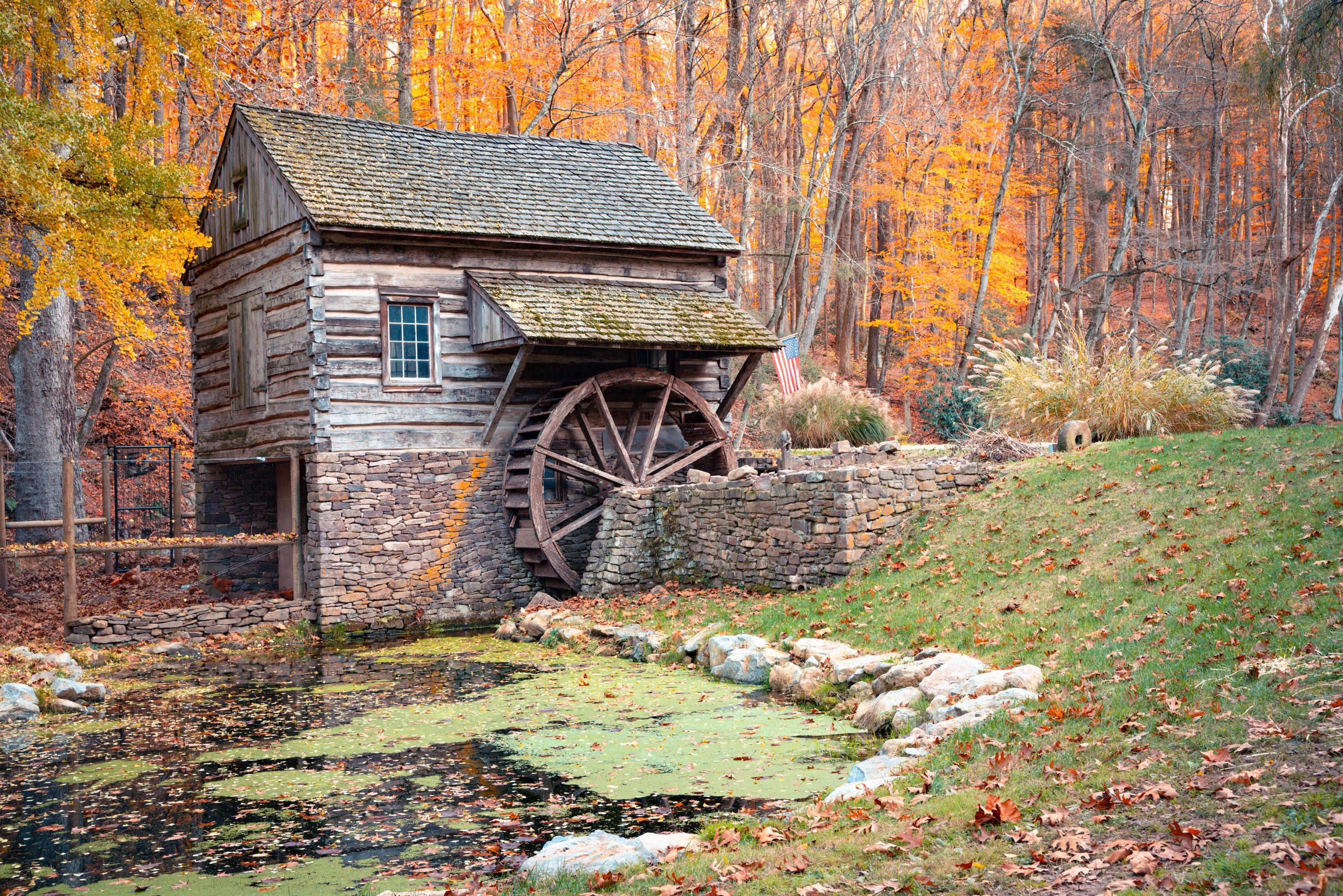 A wooden mill with a water wheel, surrounded by autumn trees and a small pond.