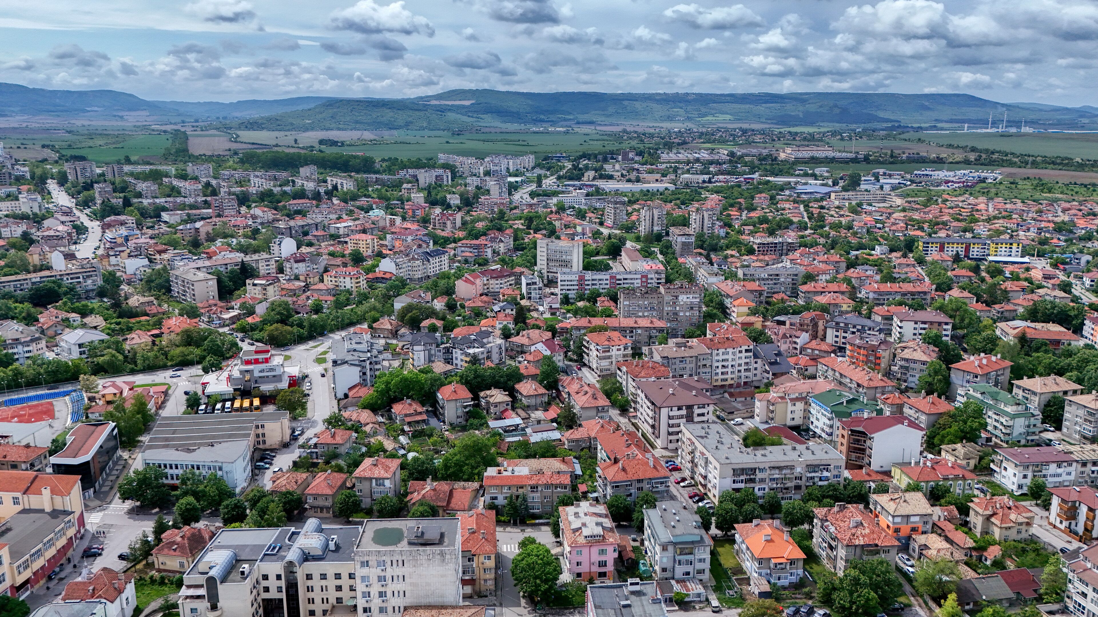 A densely populated urban area with a mix of residential and commercial buildings, surrounded by green hills and a cloudy sky.