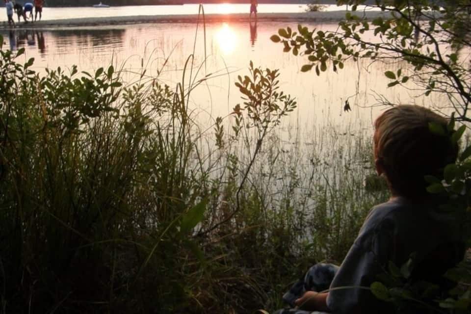 A person sitting by a lake at sunset with trees and a boat in the background.