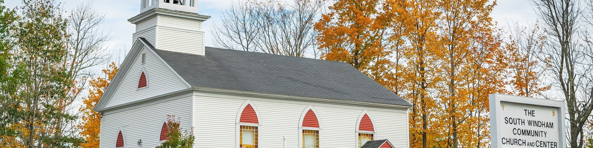 A white church with a steeple and red accents, surrounded by autumn trees.