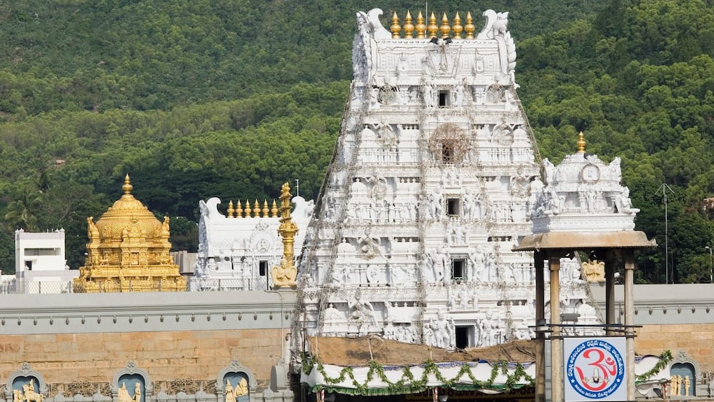 Ein weißer Tempel mit aufwendigen Schnitzereien und goldenen Akzenten, umgeben von üppigen, grünen Hügeln.