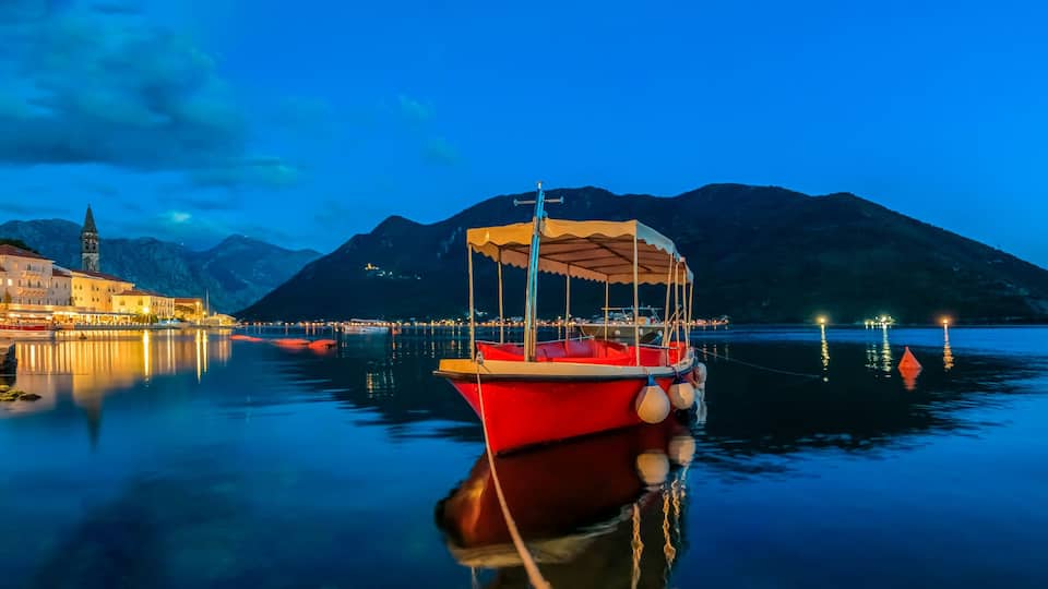 A calm lake at dusk with a red boat anchored near the shore, a church with a bell tower, and mountains in the background.