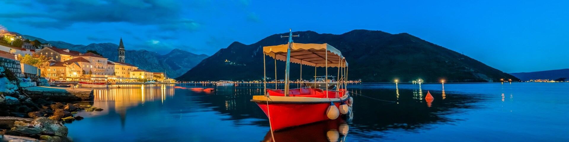 Un lago tranquilo al anochecer con un barco rojo anclado cerca de la orilla, una iglesia con un campanario y montañas al fondo.