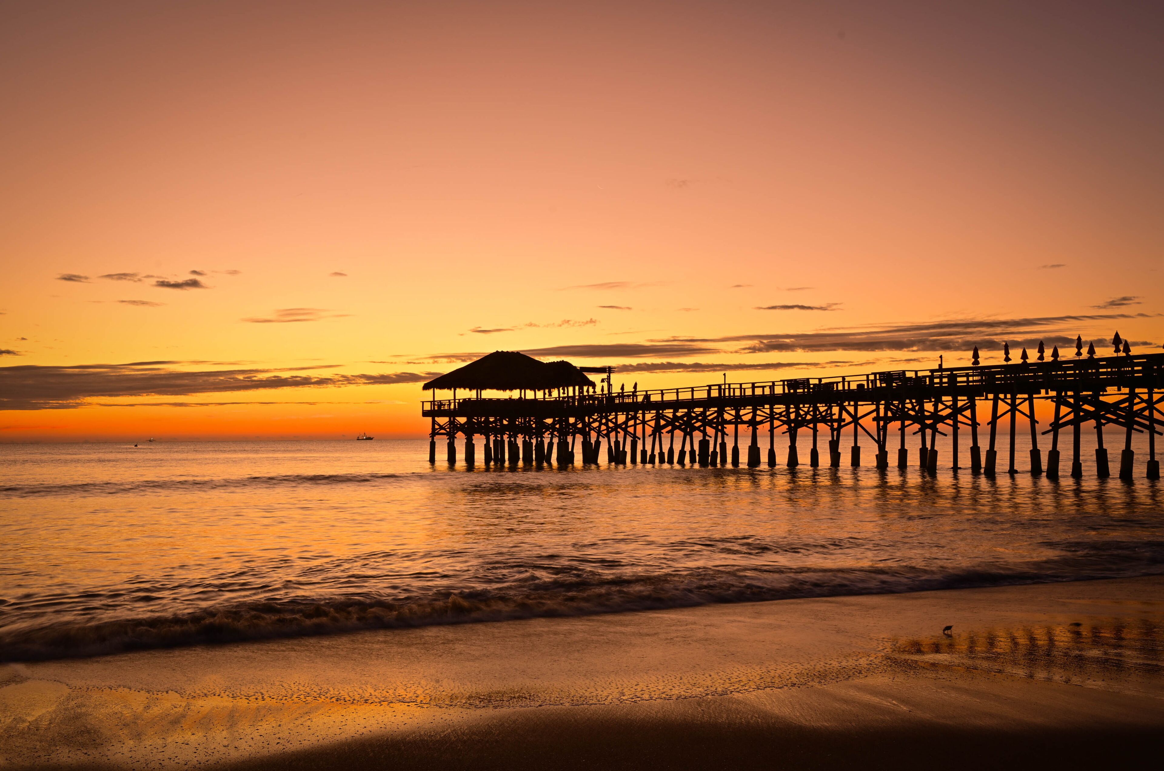 A pier extending into the ocean at sunset with a beach in the foreground.