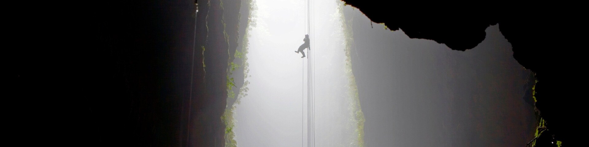 Una persona haciendo rappel por un acantilado escarpado y rocoso, con la luz del sol entrando por una abertura estrecha en la parte superior.