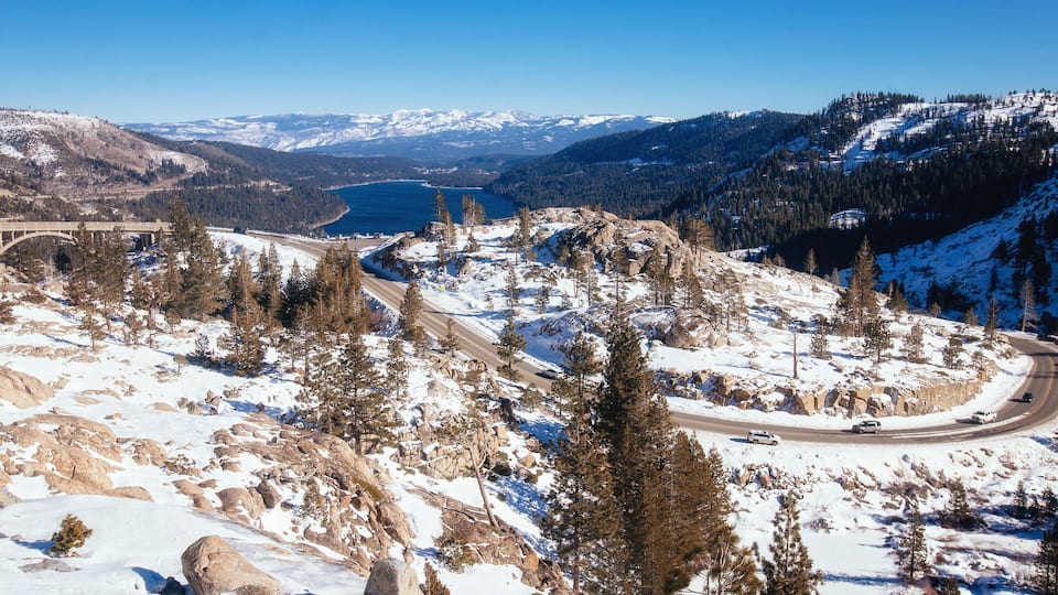 A snow-covered mountain landscape with a winding road, a bridge, and a lake in the background.
