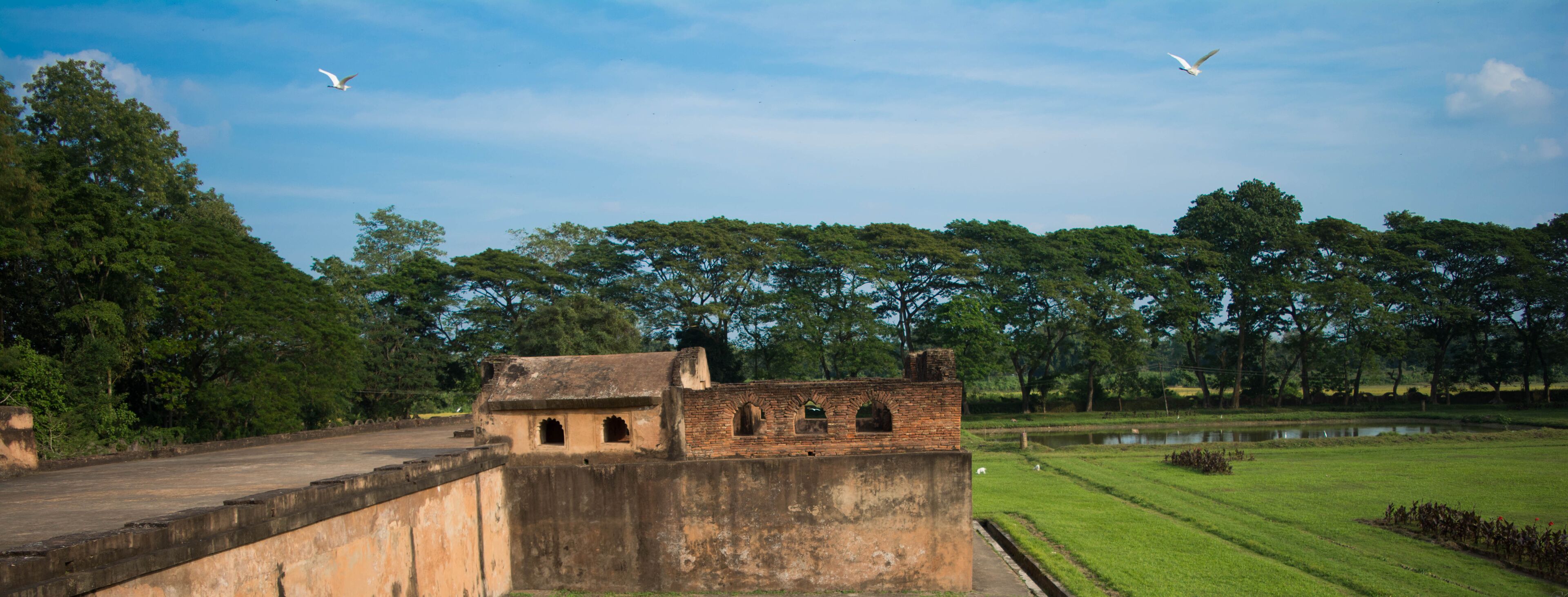 A historic fort with arched windows, a grassy field, a pond, and trees in the background.