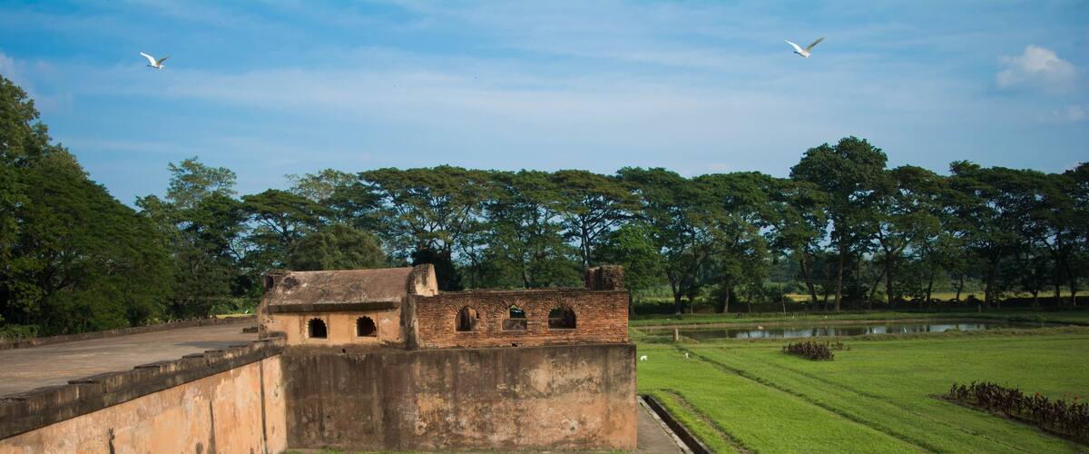 A historic fort with arched windows, a grassy field, a pond, and trees in the background.