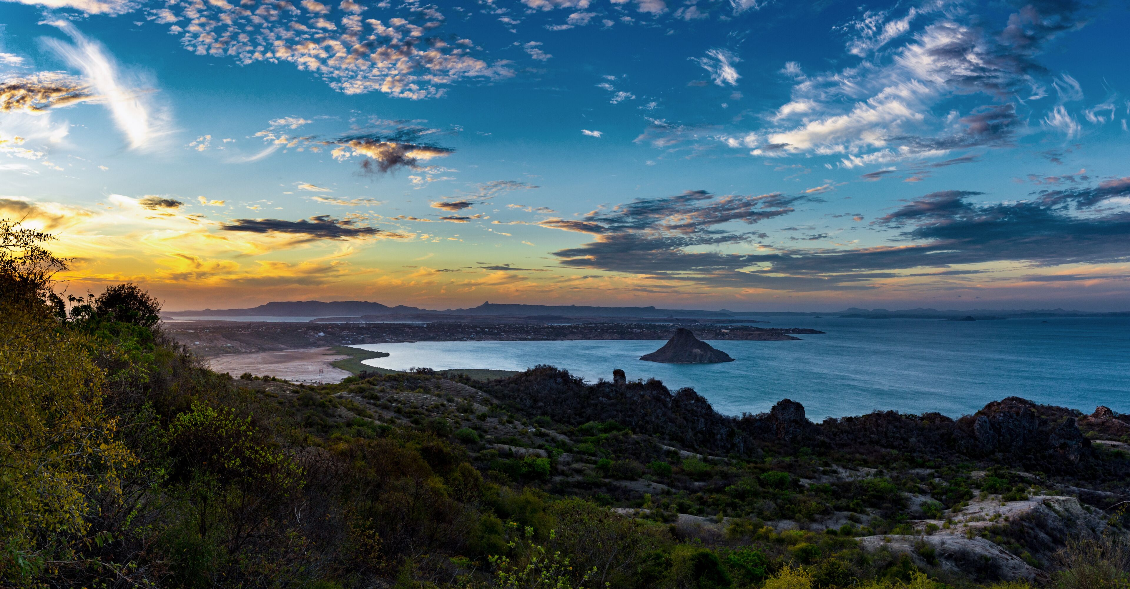 A coastal landscape with a prominent rock formation, a beach, and a distant city skyline.
