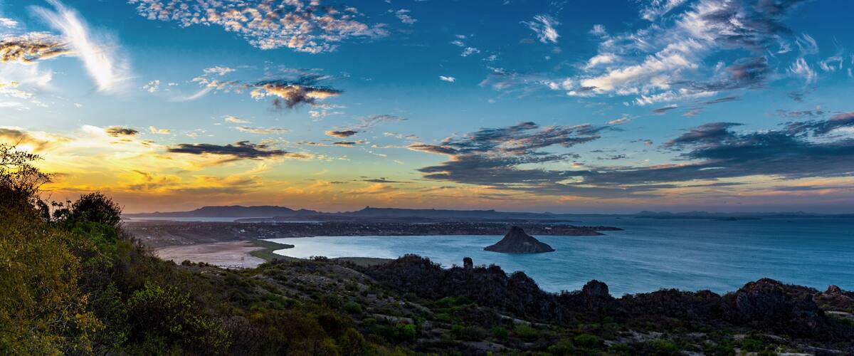 A coastal landscape with a prominent rock formation, a beach, and a distant city skyline.