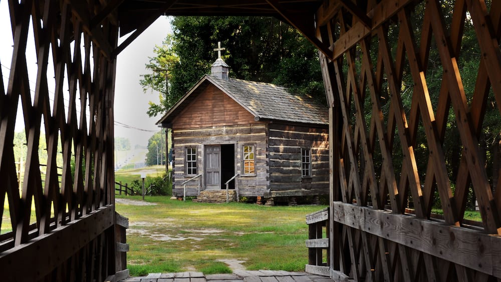 A rustic wooden cabin with a cross on the roof, surrounded by a wooden fence and a grassy yard.