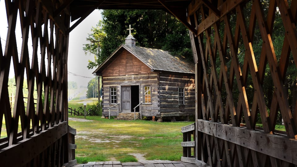 A rustic wooden cabin with a cross on the roof, surrounded by a wooden fence and a grassy yard.