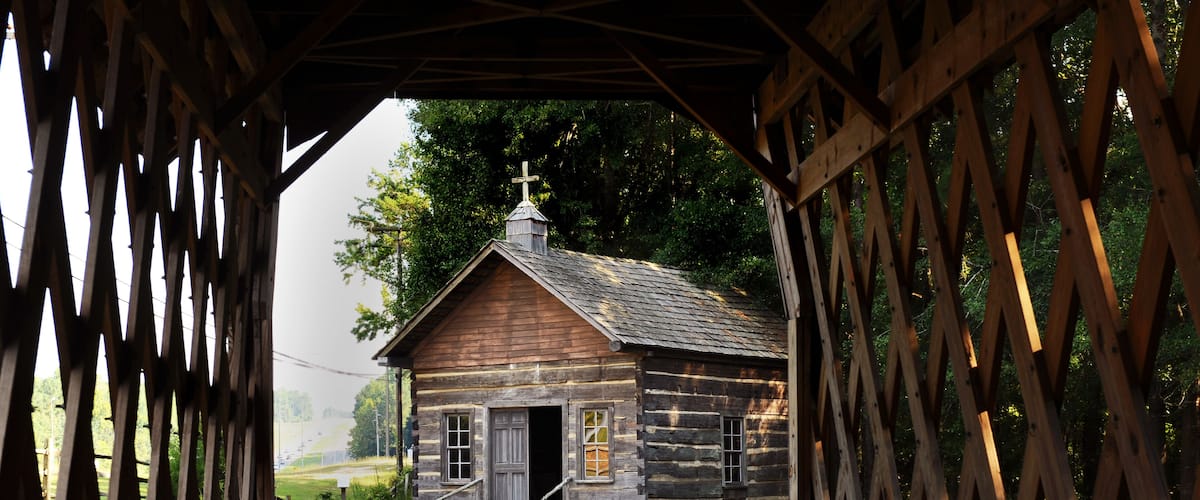 A rustic wooden cabin with a cross on the roof, surrounded by a wooden fence and a grassy yard.