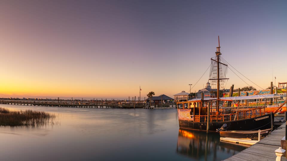 A historic sailing ship docked at a pier with a sunset in the background.