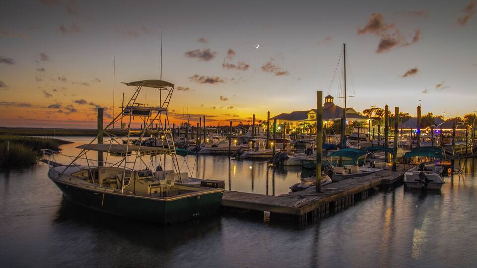 A marina at sunset with boats docked, a pier, and a building in the background.