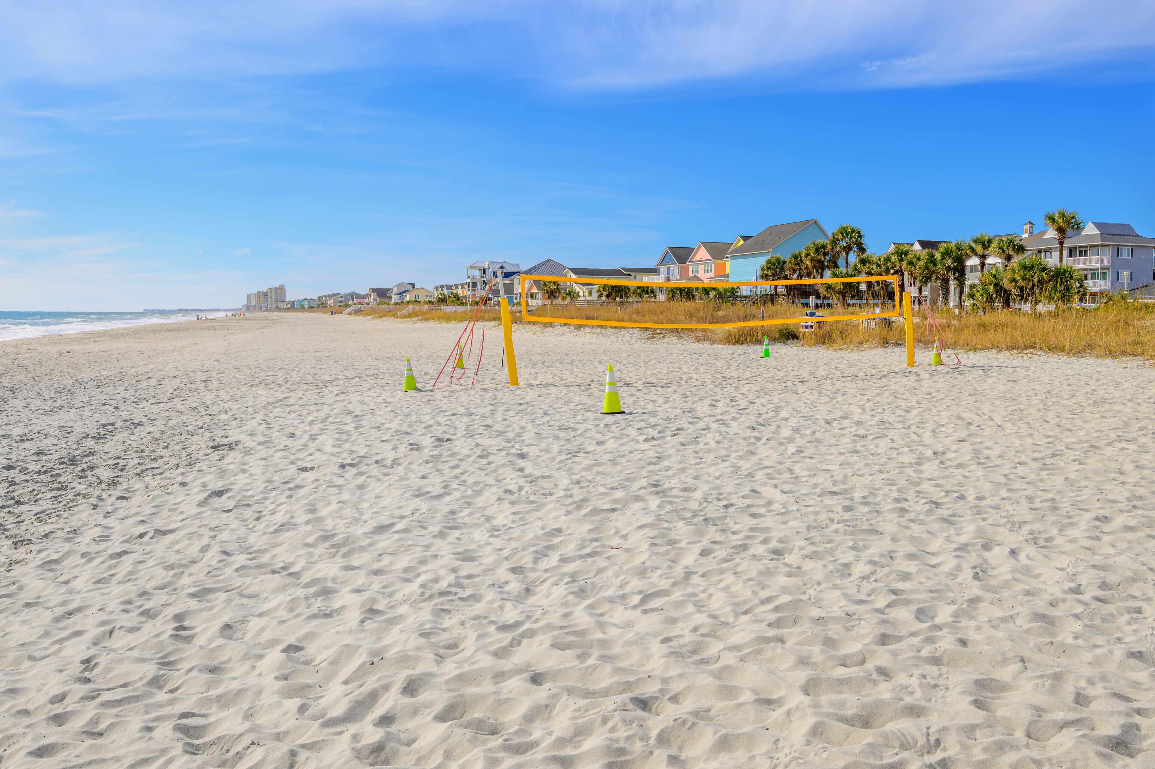 The wide sandy beach and dry sea grass along the Grand Strand Atlantic coastline, with colorful vacation homes along the shoreline and a volleyball net on the dunes, in Surfside Beach, South Carolina.