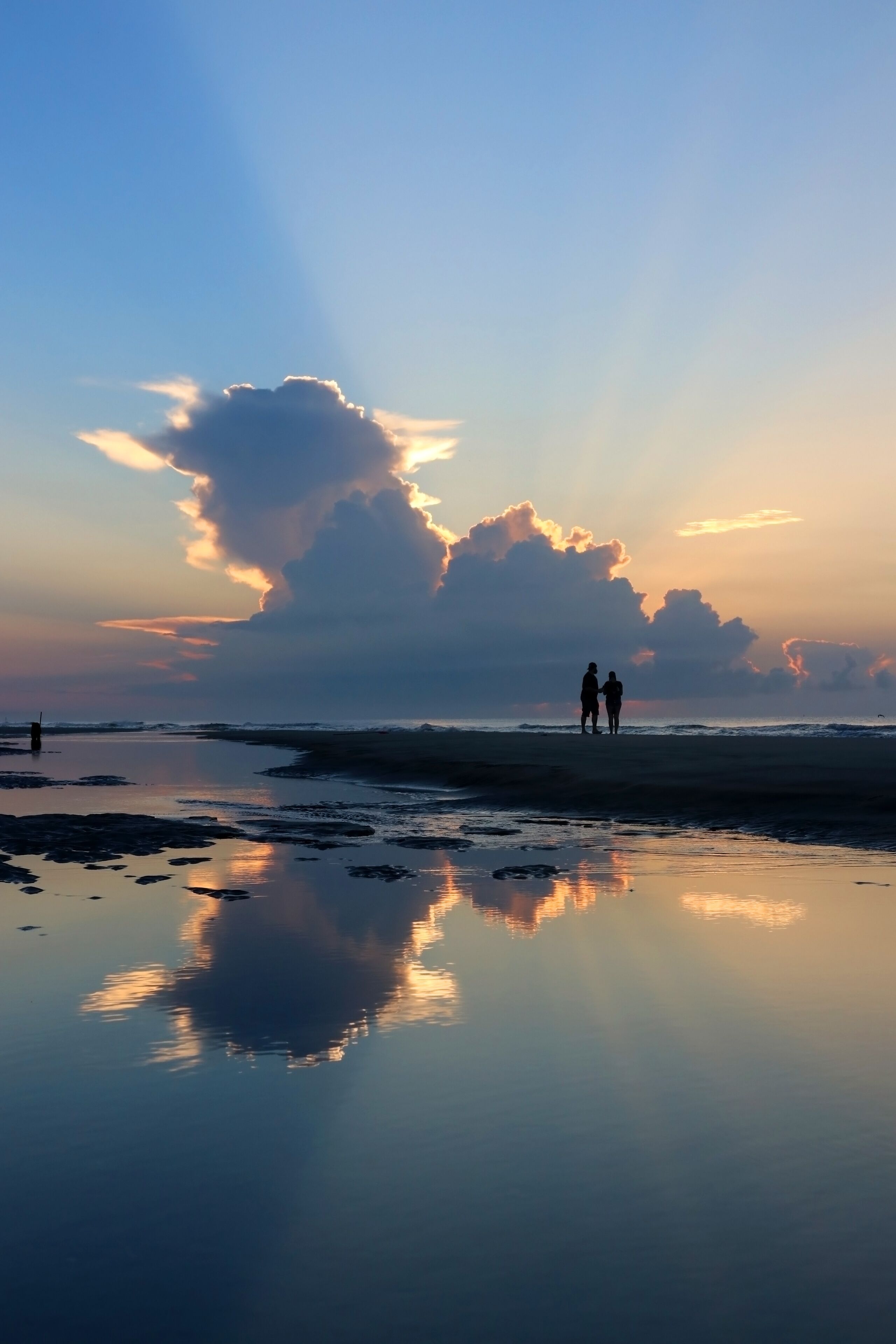 A beach with calm water reflecting clouds and two people walking.