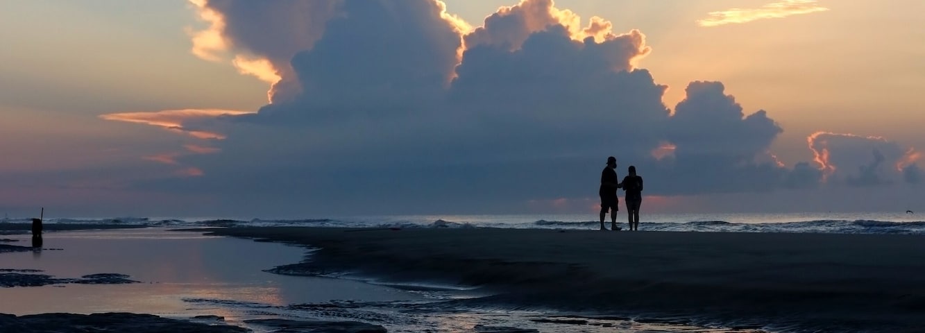 Una playa con agua tranquila que refleja nubes y dos personas caminando.