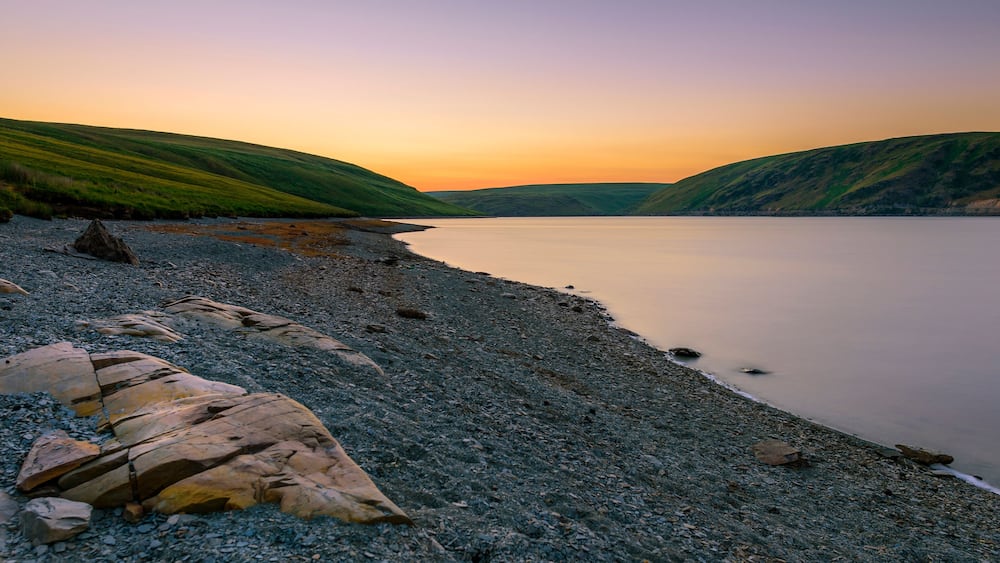 A pebbly beach with large rocks and hills in the background during sunset.