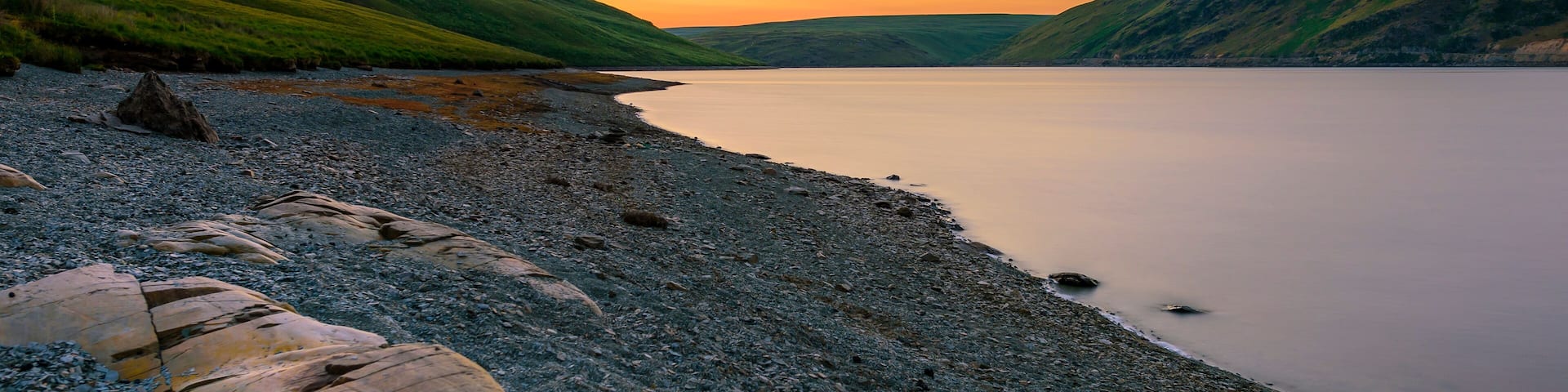 A pebbly beach with large rocks and hills in the background during sunset.