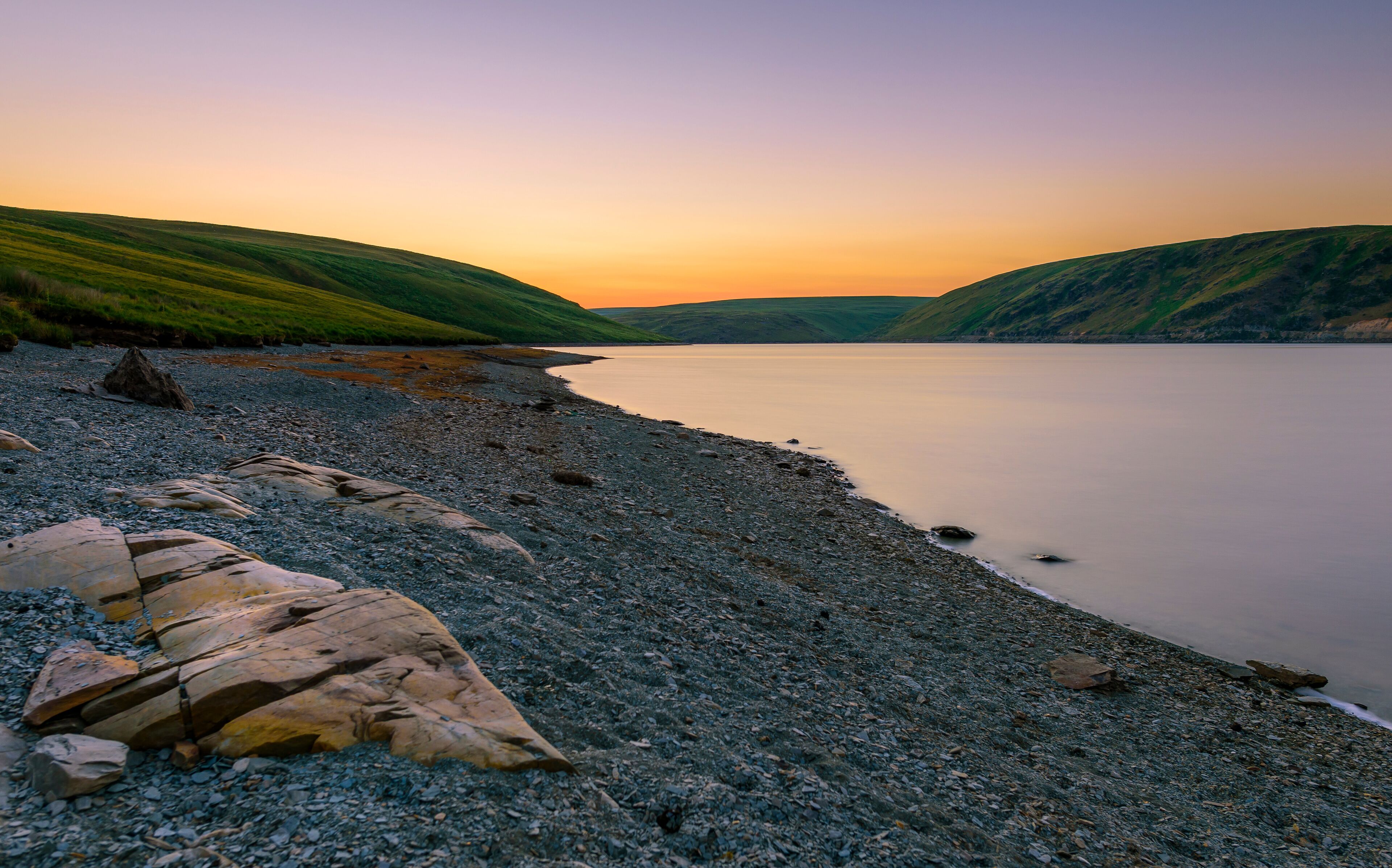 A pebbly beach with large rocks and hills in the background during sunset.