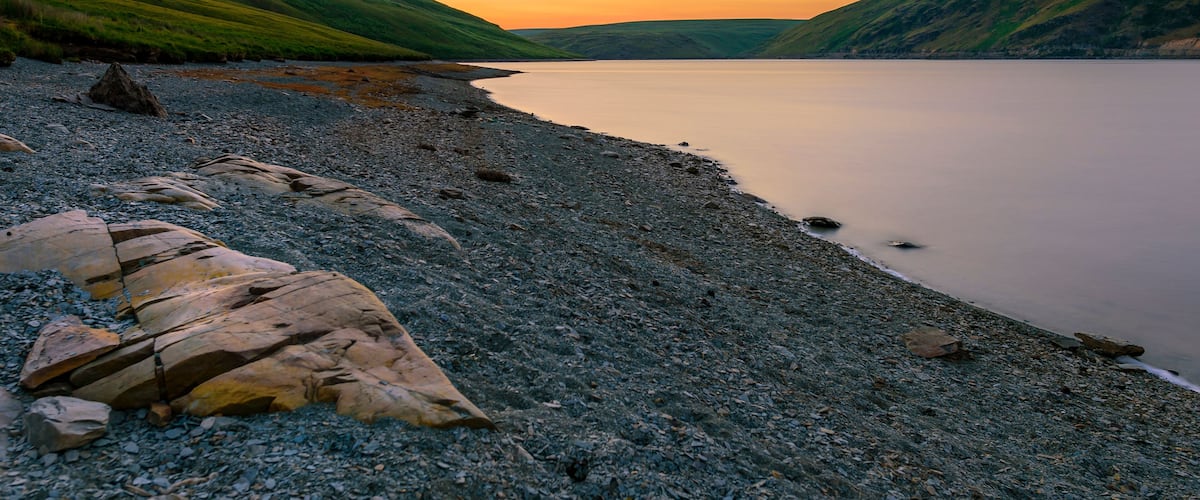 A pebbly beach with large rocks and hills in the background during sunset.