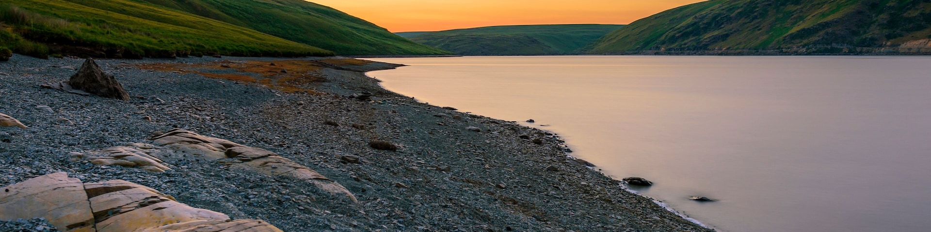 A pebbly beach with large rocks and hills in the background during sunset.