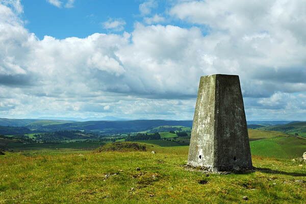 A solitary stone monument in a grassy field with rolling hills in the background under a partly cloudy sky.
