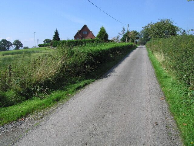 A narrow rural road flanked by greenery and a red house in the distance.