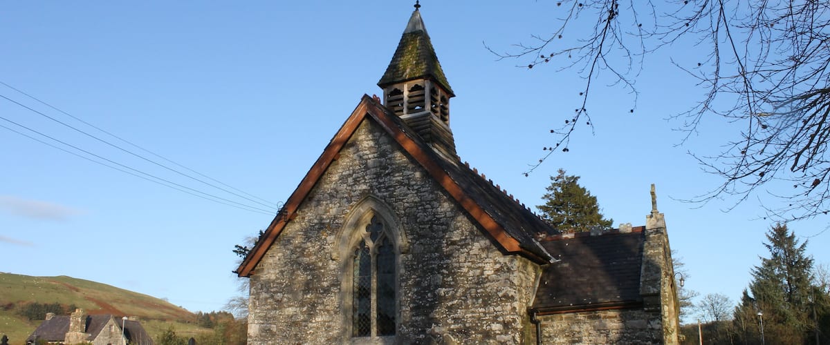 A stone church with a pointed steeple and a clock tower, surrounded by a cemetery with headstones.