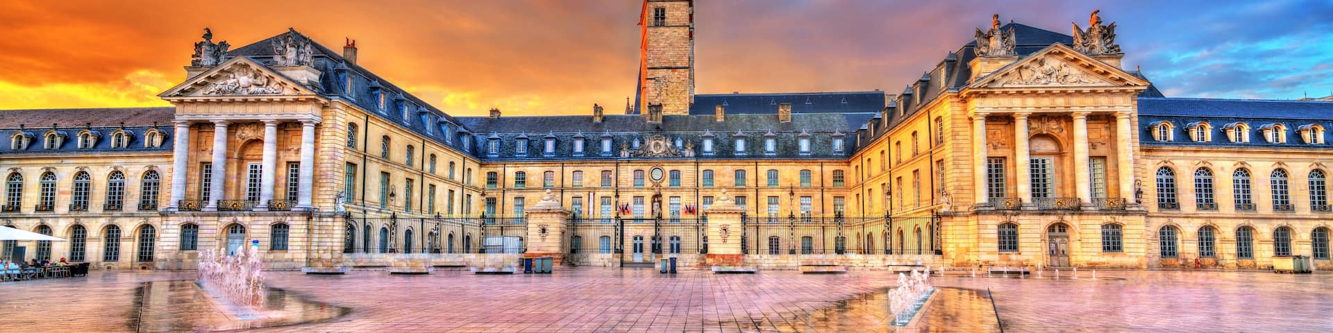A grand historic building with a central tower, surrounded by a spacious courtyard with a fountain.