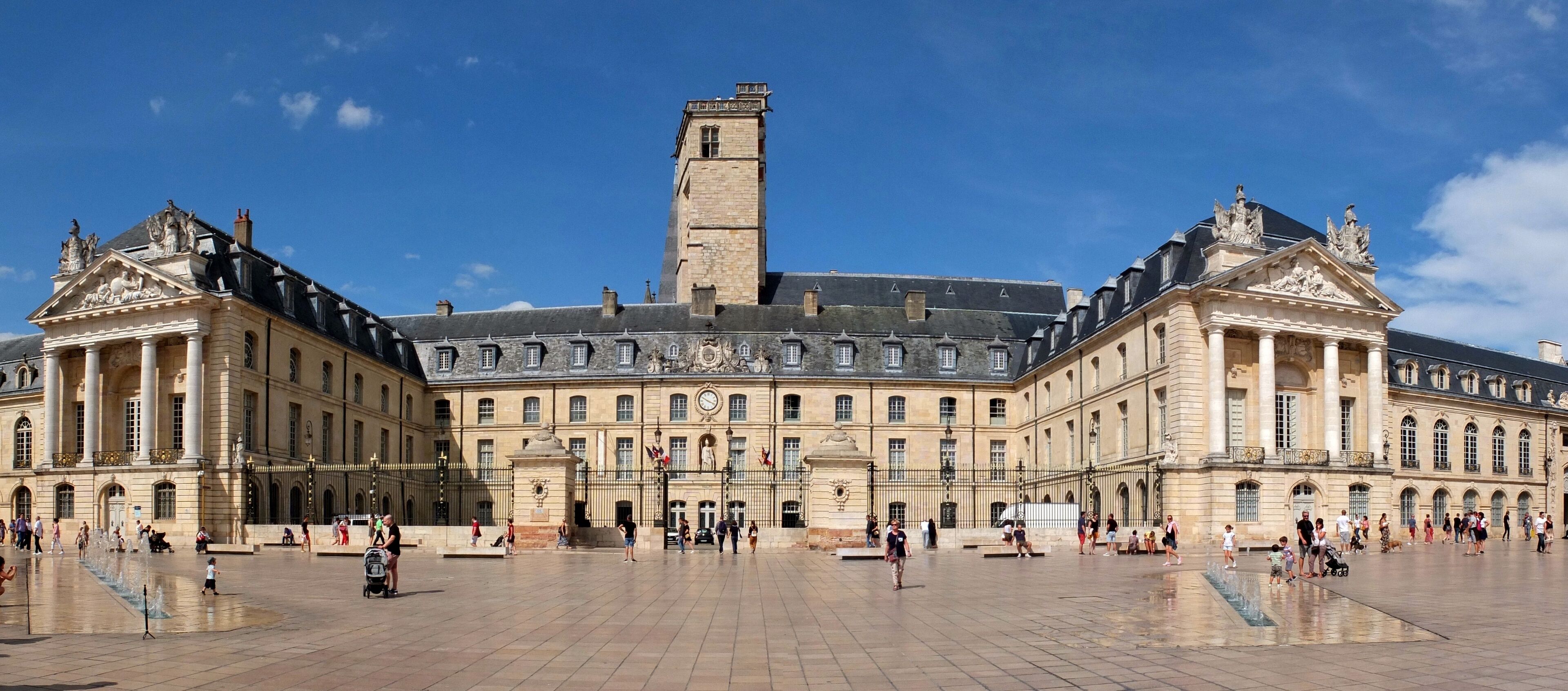 A large historic building with a central tower, surrounded by a spacious courtyard with people walking and a fountain.