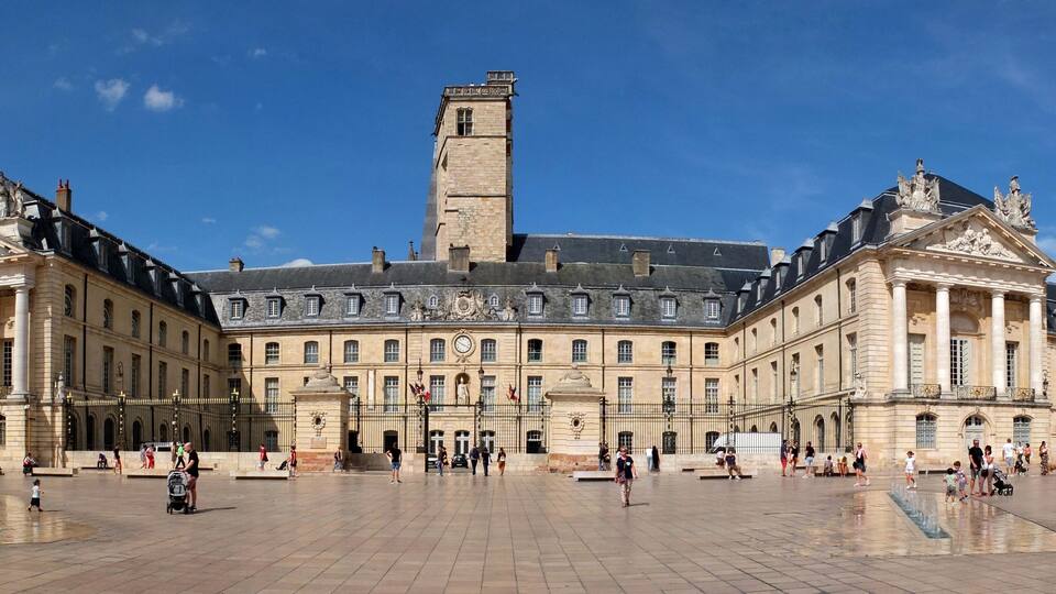 A large historic building with a central tower, surrounded by a spacious courtyard with people walking and a fountain.