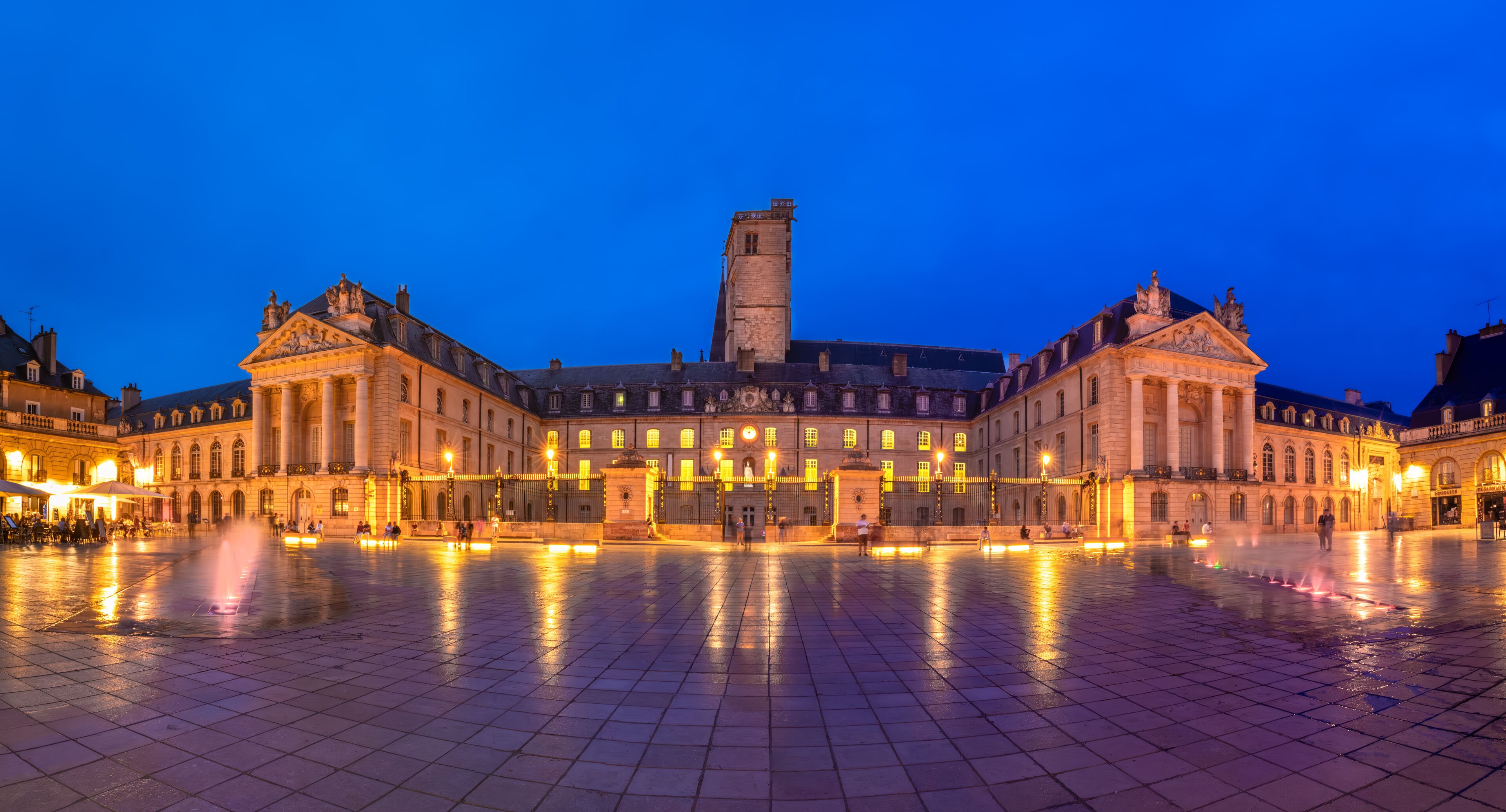 A historic square with a central fountain, ornate buildings, and a tall tower.