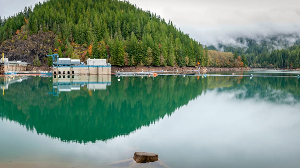 A calm lake reflecting a forested mountain, a bridge, and industrial structures.