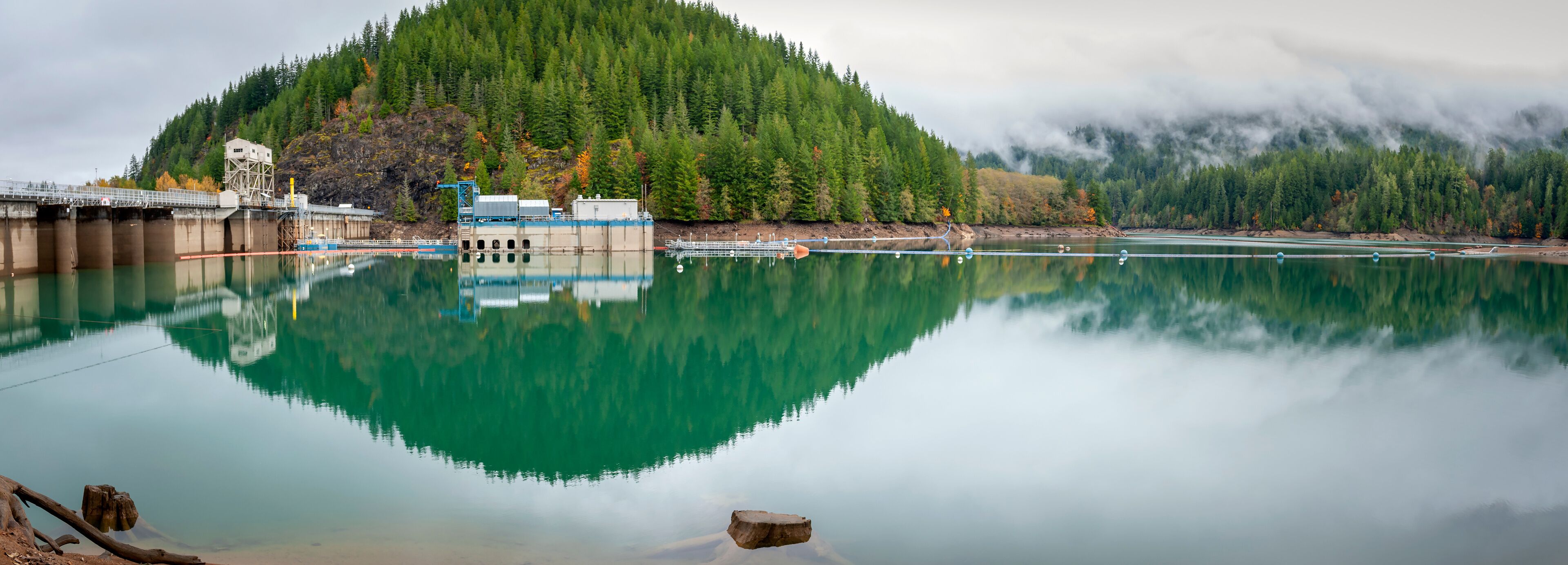 A calm lake reflecting a forested mountain, a bridge, and industrial structures.
