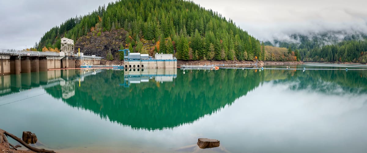 A calm lake reflecting a forested mountain, a bridge, and industrial structures.