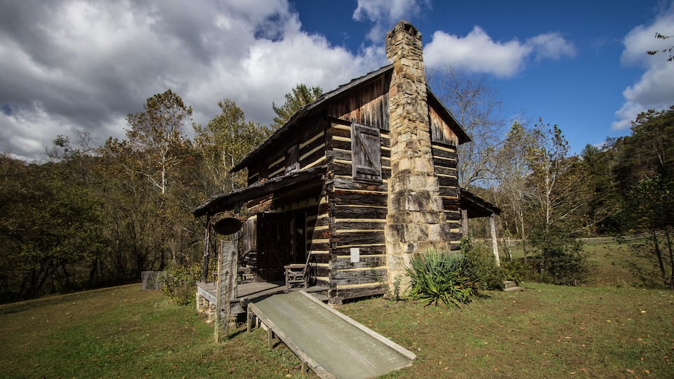 A rustic log cabin with a stone chimney, surrounded by a grassy yard and trees.