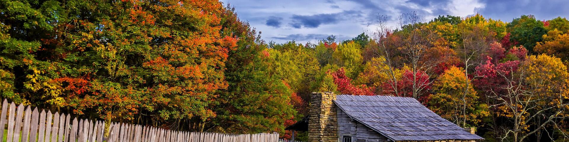 A rustic stone house surrounded by a wooden fence, set against a backdrop of colorful autumn trees.