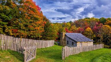 A rustic stone house surrounded by a wooden fence, set against a backdrop of colorful autumn trees.