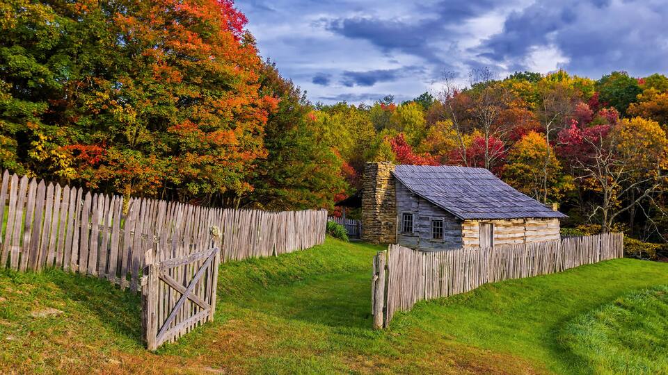 A rustic stone house surrounded by a wooden fence, set against a backdrop of colorful autumn trees.
