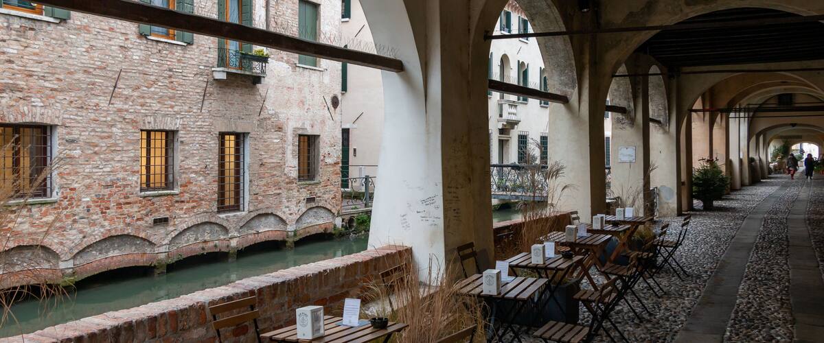 A riverside café with outdoor seating, brick buildings, and arched walkways.