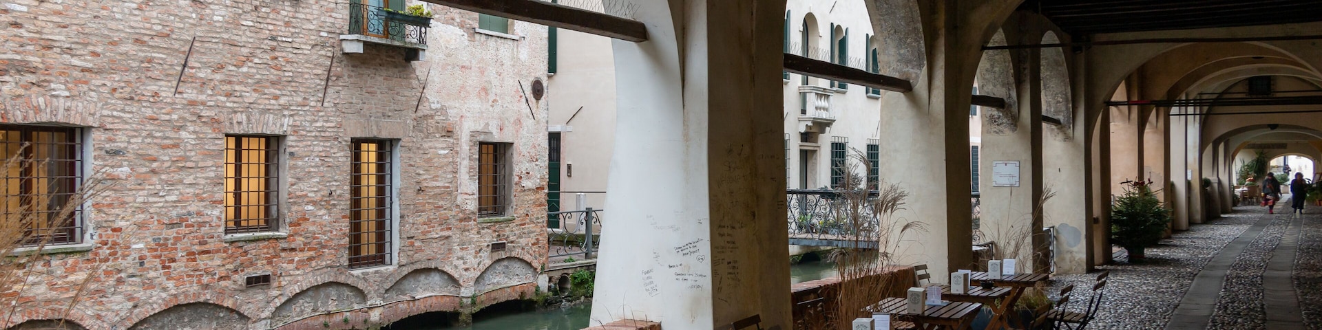A riverside café with outdoor seating, brick buildings, and arched walkways.