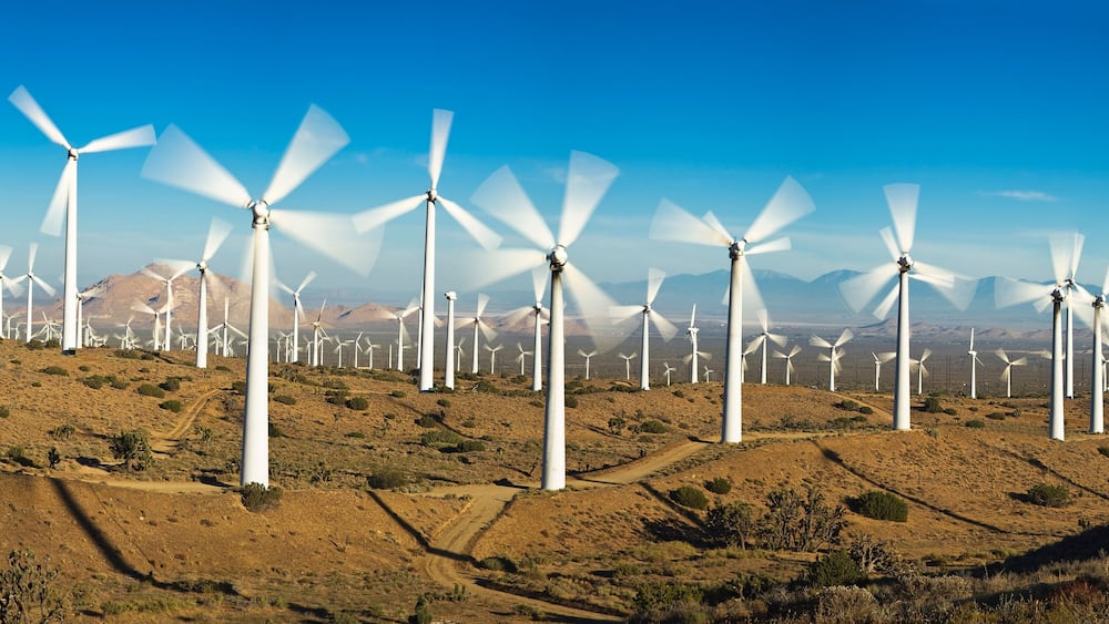 Ein Windpark mit zahlreichen weißen Windturbinen in hügeliger Landschaft unter blauem Himmel.