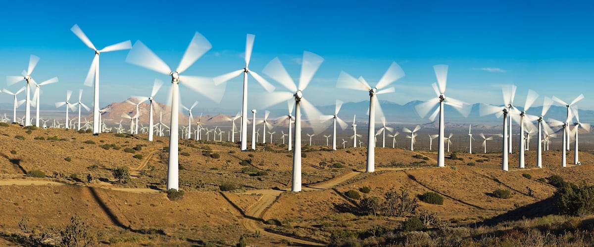 Ein Windpark mit zahlreichen weißen Windturbinen in hügeliger Landschaft unter blauem Himmel.